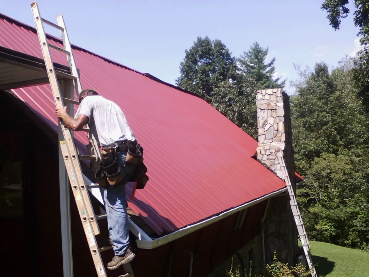 Roofer on a ladder beside a red metal roof and stone chimney — Soffit & Fascia Repair by Roofing Brothers in Short Hills